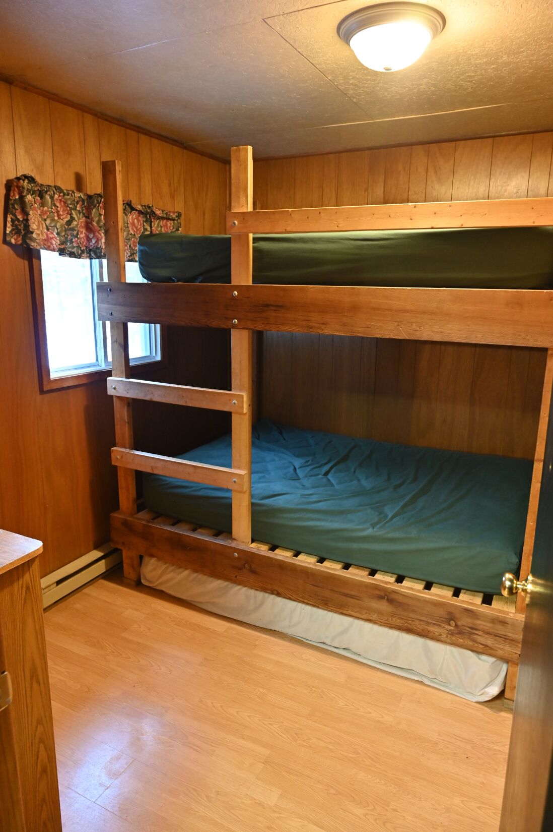 A bunk bed with a wooden frame and green bedding, a window with floral curtains, and a wooden floor.