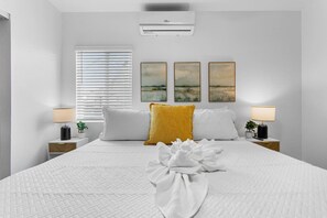 A hotel room with a large bed, white bedding, two bedside lamps, a window with blinds, and three wall-mounted paintings.