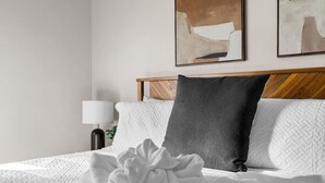 A neatly made bed with a white duvet, a dark pillow, and a large white decorative pillow. There is a lamp on a bedside table and two abstract paintings on the wall. - Modern 5 BD Home w/ Smart TV and Ping Pong (North Las Vegas)