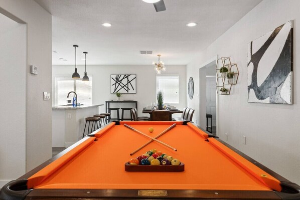 A pool table with a billiard cue and balls, a dining area with chairs and a table, and a wall with abstract art.