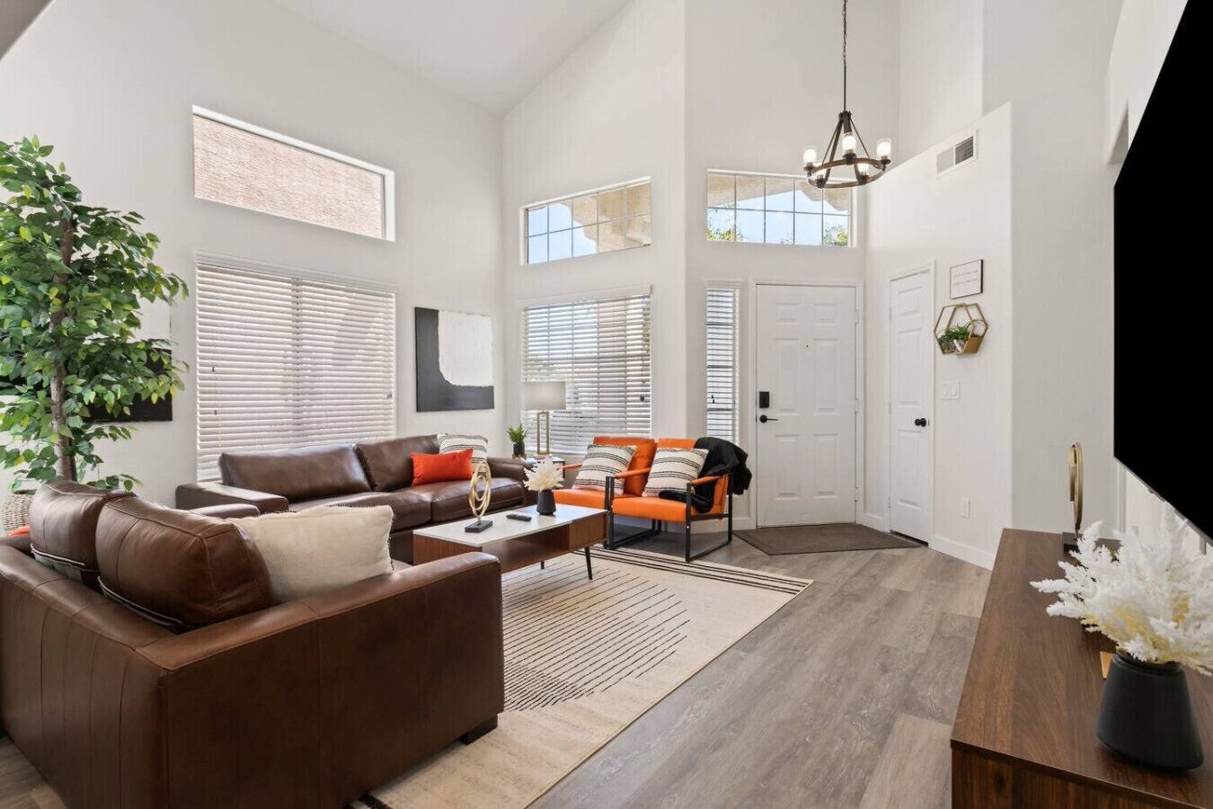 A spacious living room with a brown leather sofa, orange armchairs, a wooden coffee table, and a large window.