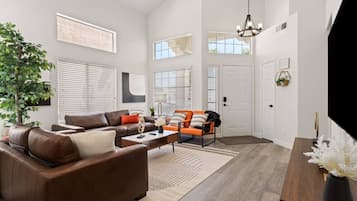 A spacious living room with a brown leather sofa, orange armchairs, a wooden coffee table, and a large window.