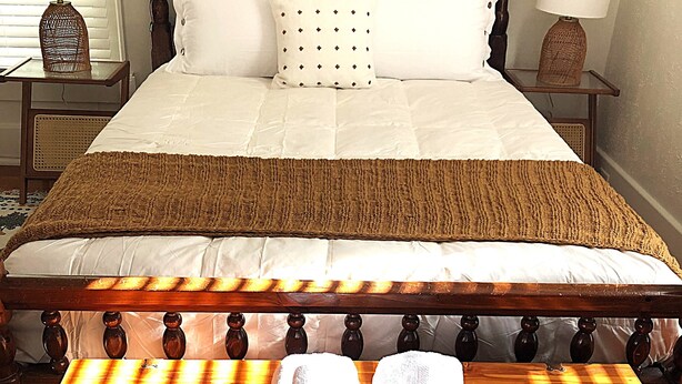 Ein ordentlich bezogenes Bett mit hölzernem Kopfteil, weißer Bettwäsche und einer braunen Tagesdecke. Auf einem Holztisch vor dem Bett liegen zwei zusammengerollte Handtücher.