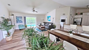 A modern living room with a sofa, dining table, and chairs, a kitchen area with a refrigerator and cabinets, and a large window with a view of greenery. - Pool & Beach – Shark Teeth Hunting – Families welcome – heated Pool (Venice)