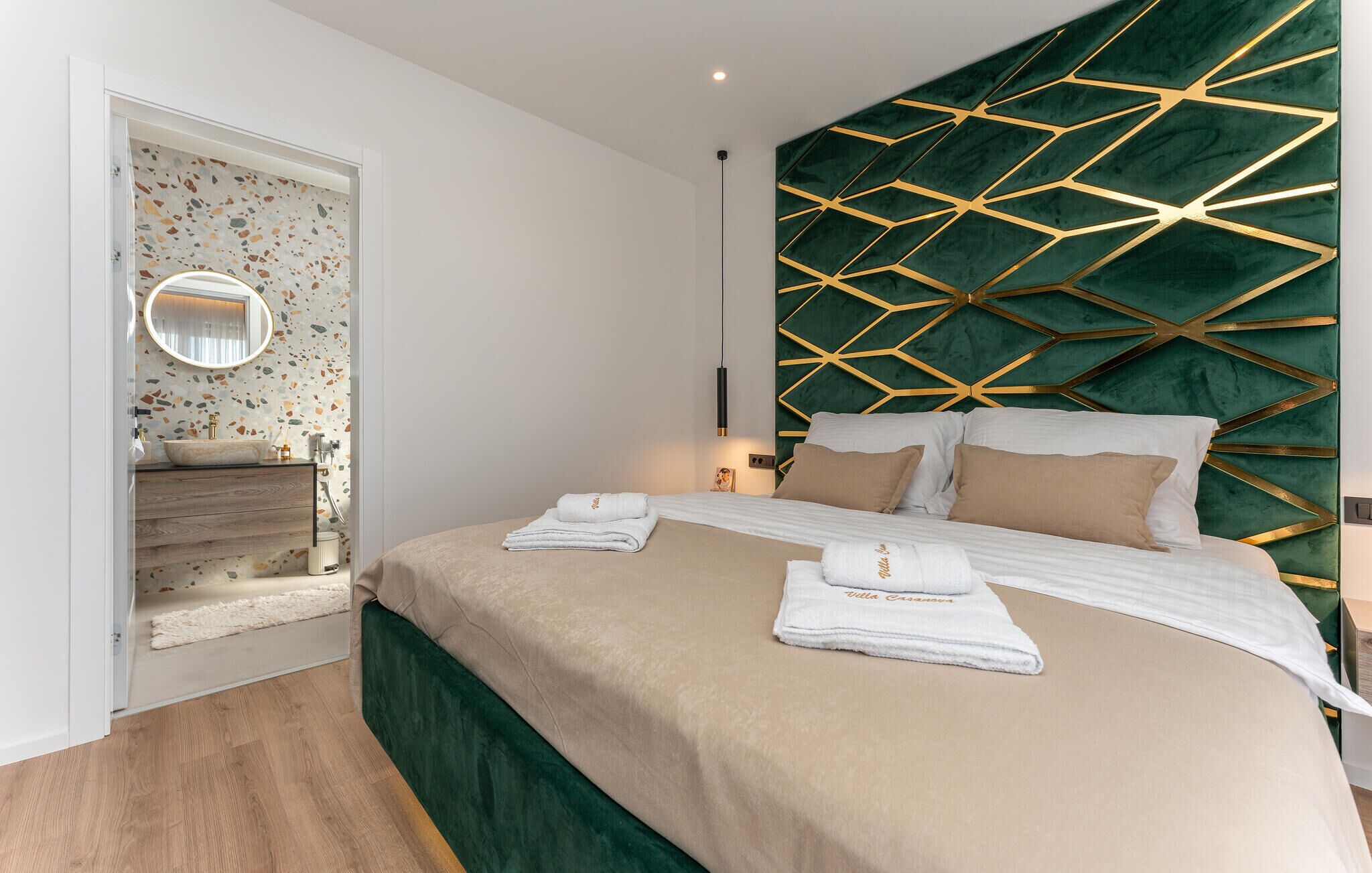 Ein modernes Schlafzimmer mit einem grünen gepolsterten Kopfteil, einem großen Bett mit weißer und beiger Bettwäsche, einem runden Spiegel über dem Waschbecken und einem Terrazzo-Boden.