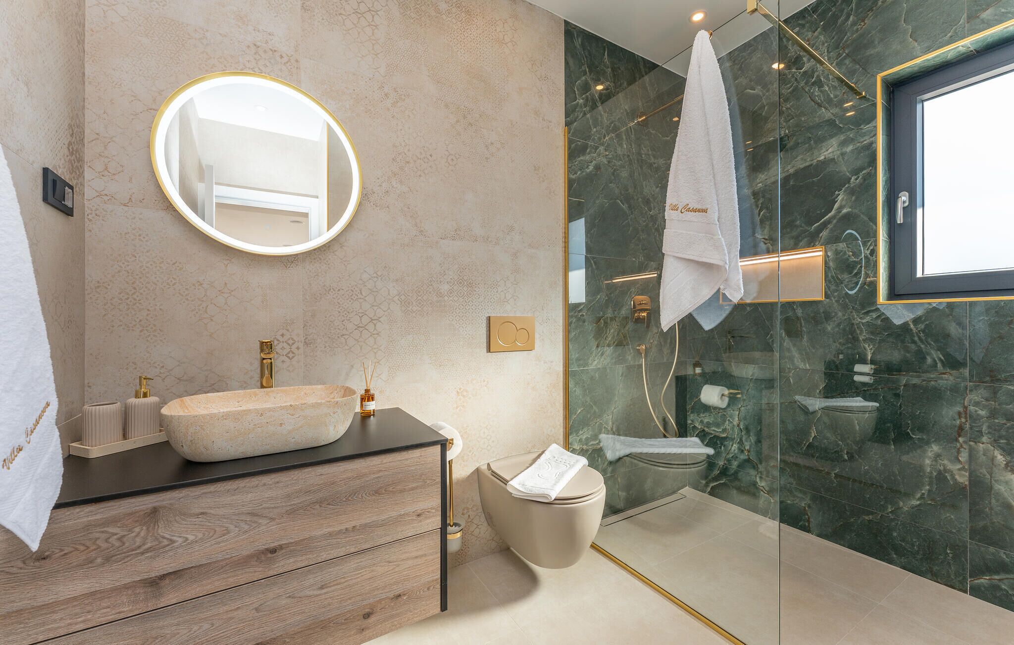 Un baño moderno con un espejo redondo, un lavabo sobre encimera, inodoro y ducha con mampara de vidrio.
