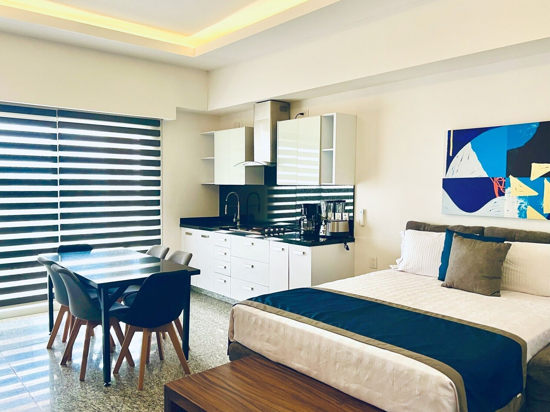 Una habitación de hotel moderna con cama, una mesa de comedor con sillas, kitchenette y un amplio ventanal con persianas.