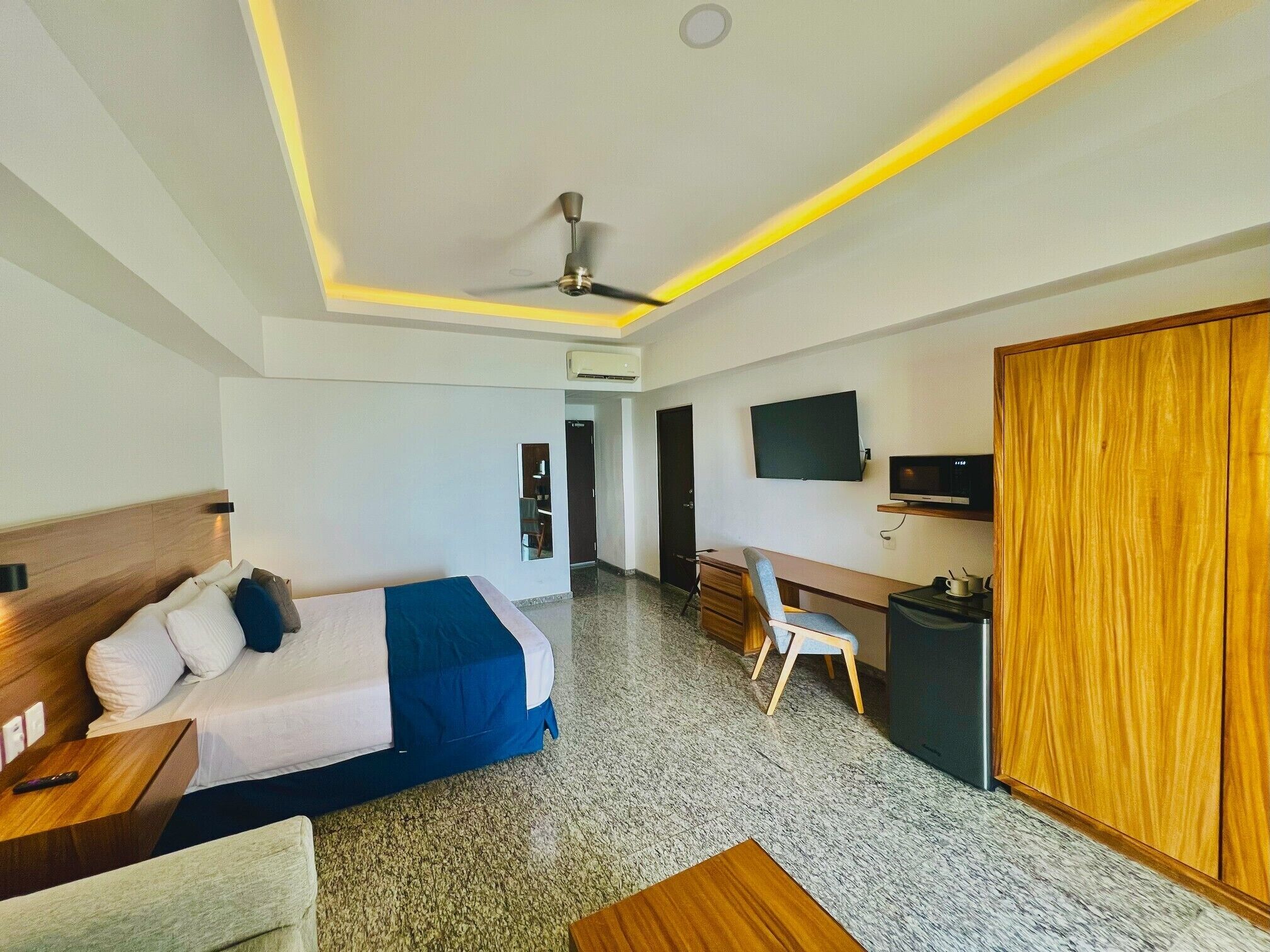 A hotel room with a bed, desk, chair, TV, wardrobe, and ceiling fan.