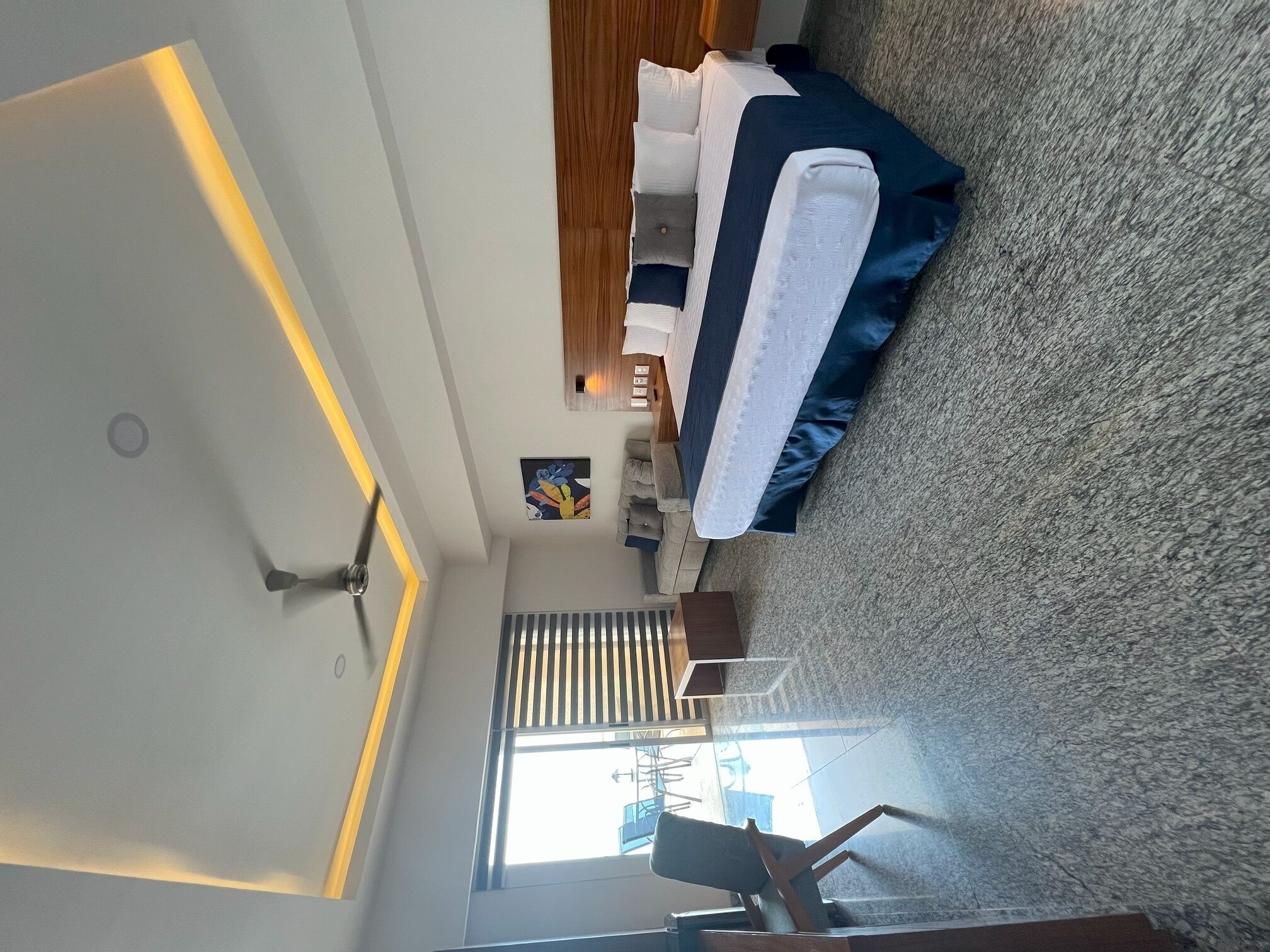 Een moderne slaapkamer met een groot bed, een houten nachtkastje, een kleine zithoek, een schilderij en een badkamer met een bad en een plafondventilator.