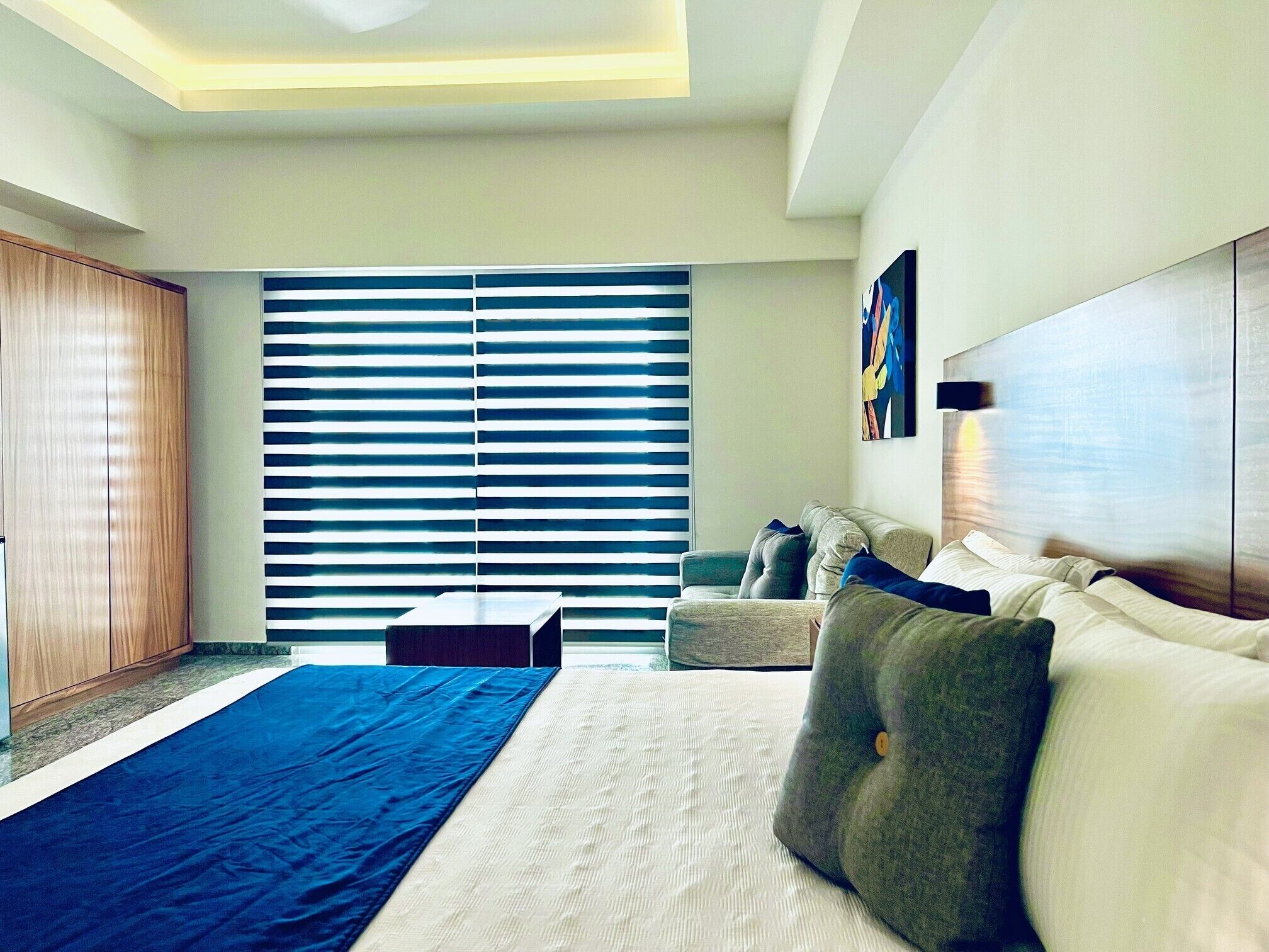 Une chambre d’hôtel moderne avec un grand lit, un canapé et un bureau. On y trouve une grande fenêtre avec des stores, un tableau au mur et un luminaire au plafond.