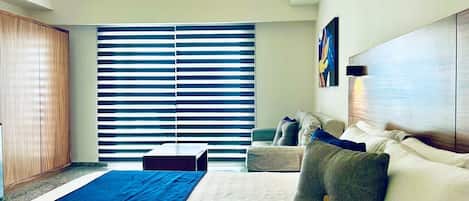 Une chambre d’hôtel moderne avec un grand lit, un canapé et un bureau. On y trouve une grande fenêtre avec des stores, un tableau au mur et un luminaire au plafond.