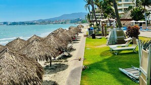 Una spiaggia con ombrelloni di paglia, palme e un albergo sullo sfondo.