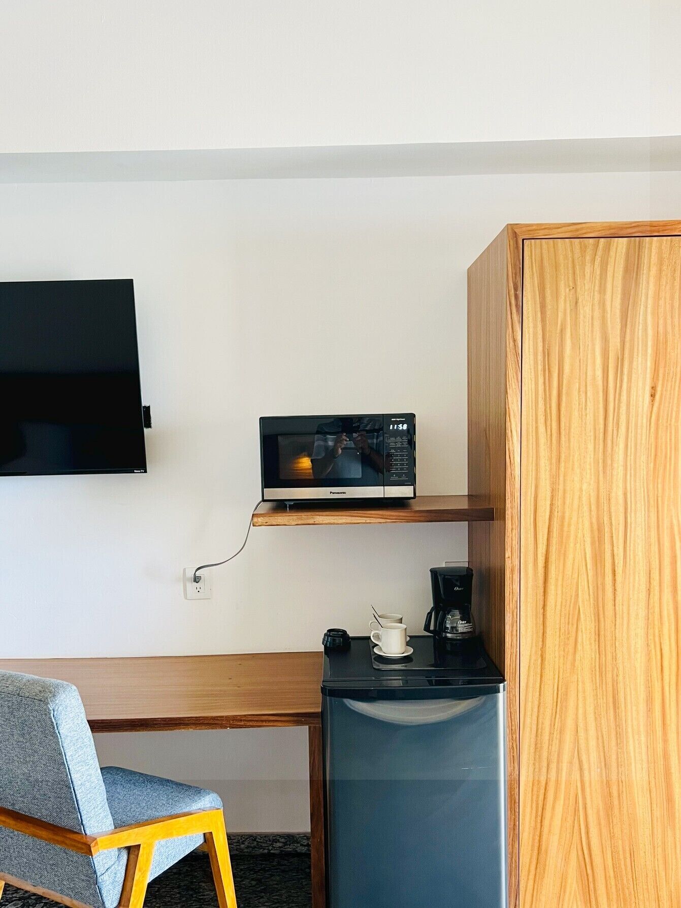 A hotel room with a wooden desk, a television mounted on the wall, a microwave on a shelf, a coffee maker, and a refrigerator.