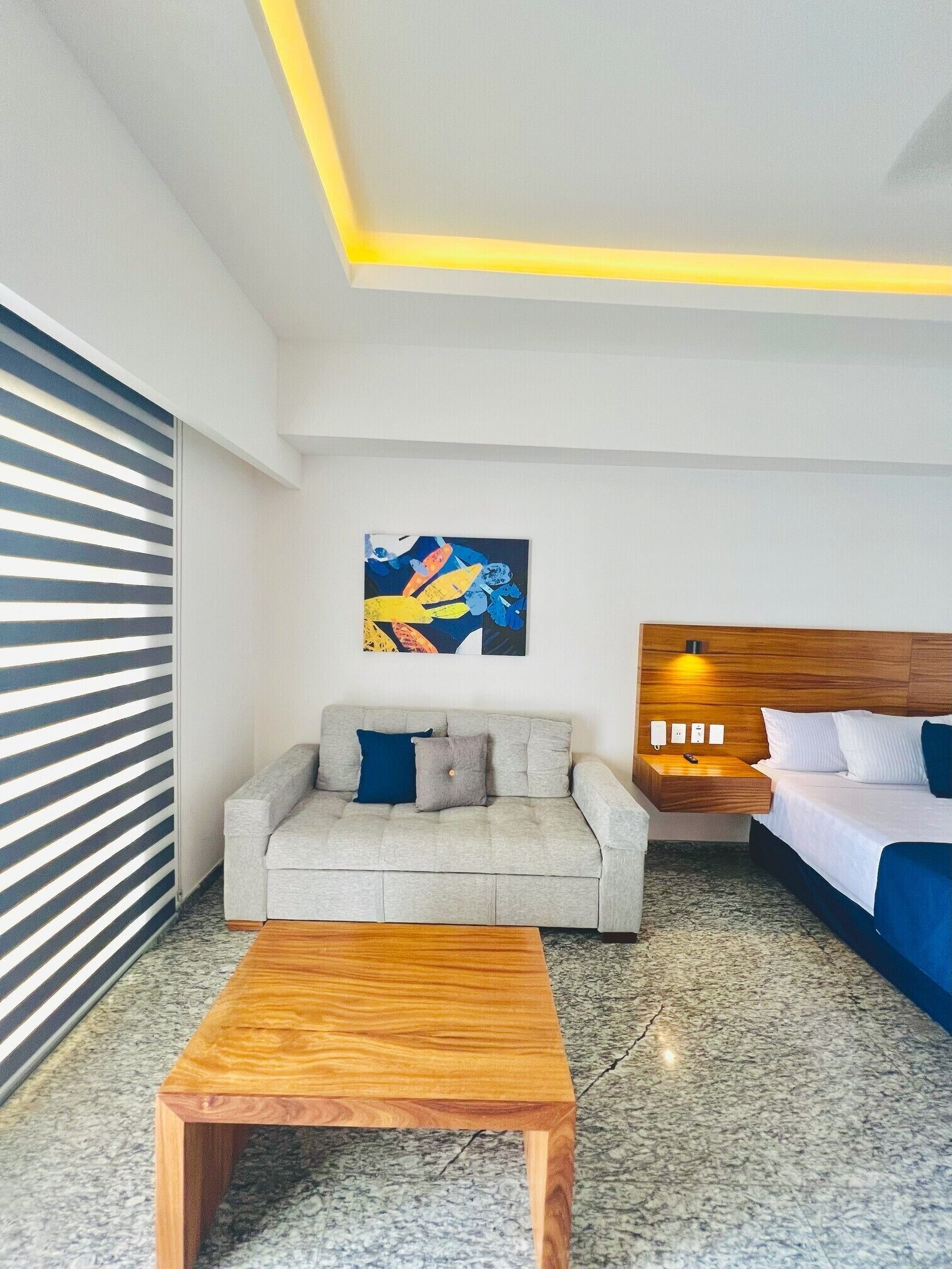 A modern hotel room with a sofa, a wooden coffee table, a bed with a headboard, a painting on the wall, and a nightstand with a lamp.