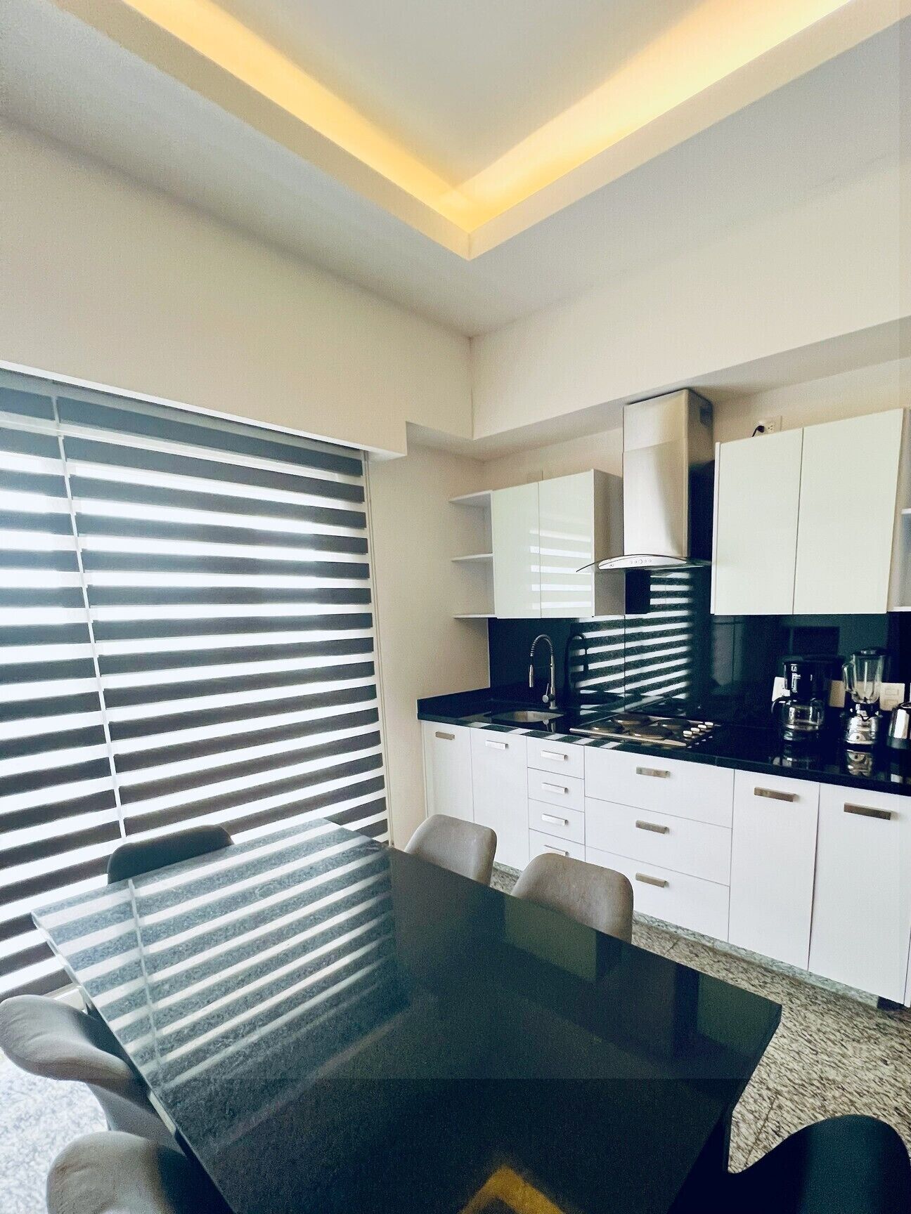 Una cocina moderna con armarios blancos, encimera negra y estufa integrada. Se observa una mesa de comedor con sillas y una ventana con persianas.
