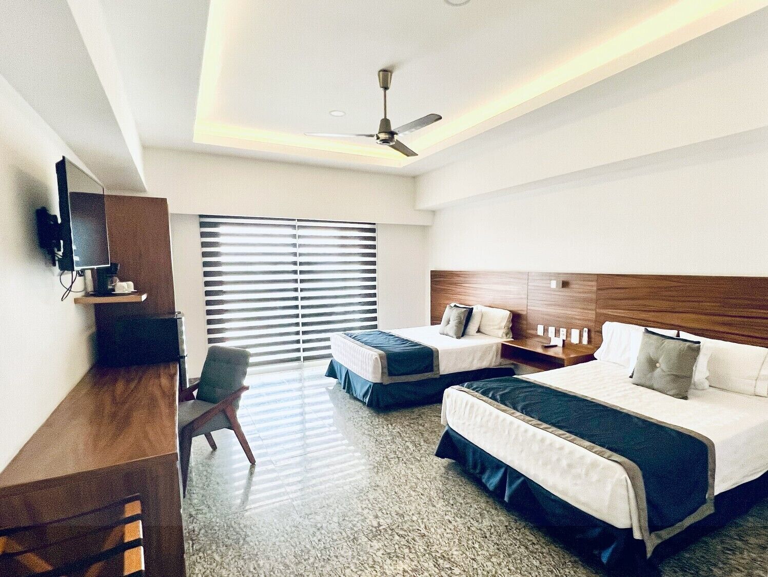 Habitación de hotel con dos camas, un escritorio con silla, televisor y ventilador de techo.