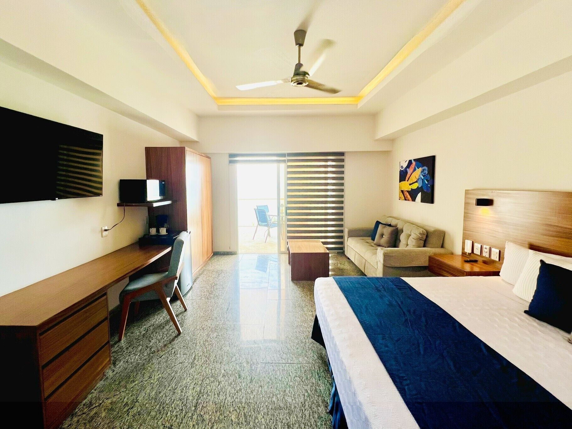 Una habitación de hotel moderna con una cama grande, un escritorio con silla, un sofá, un televisor y un ventilador de techo.