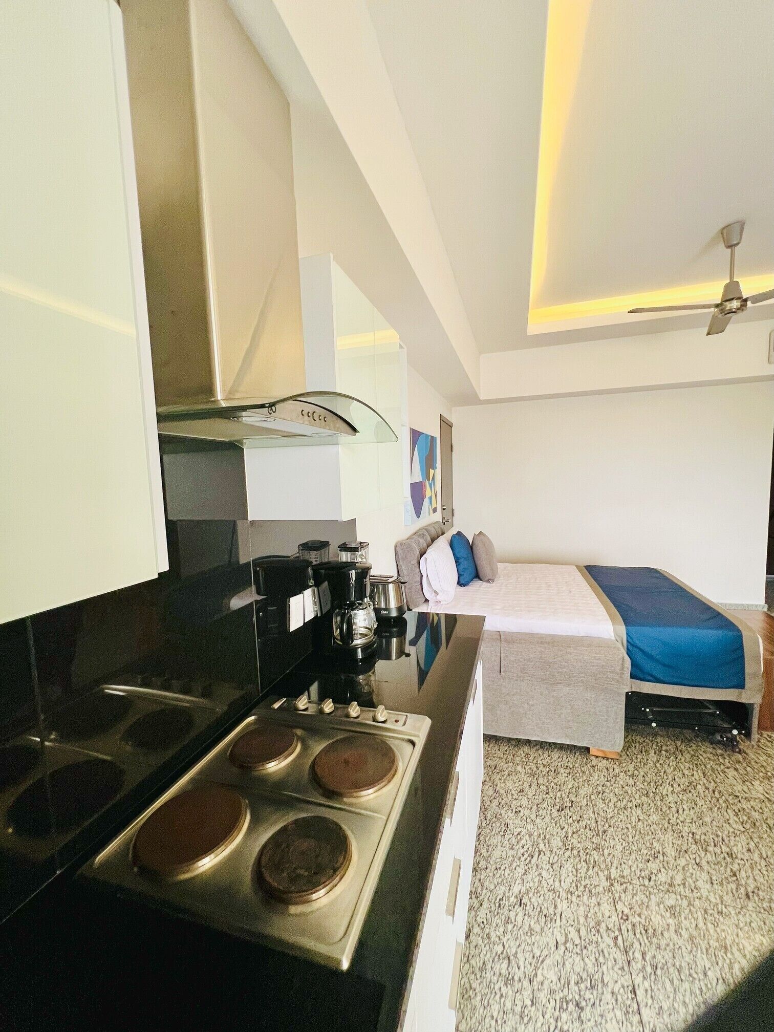 A compact hotel room with a kitchenette, a bed with a blue mattress, and a ceiling fan.