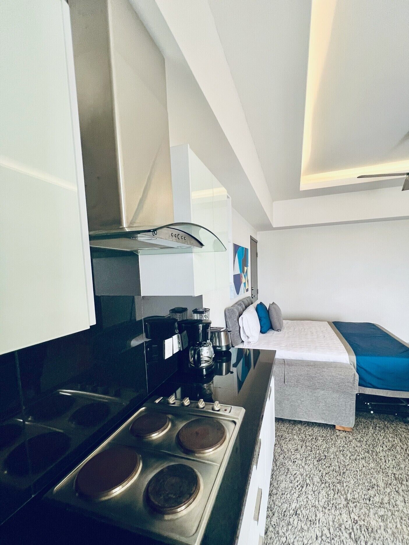 Cocina compacta con estufa, cafetera y alacenas. Se ve una cama con un edredón y almohadas en el fondo.