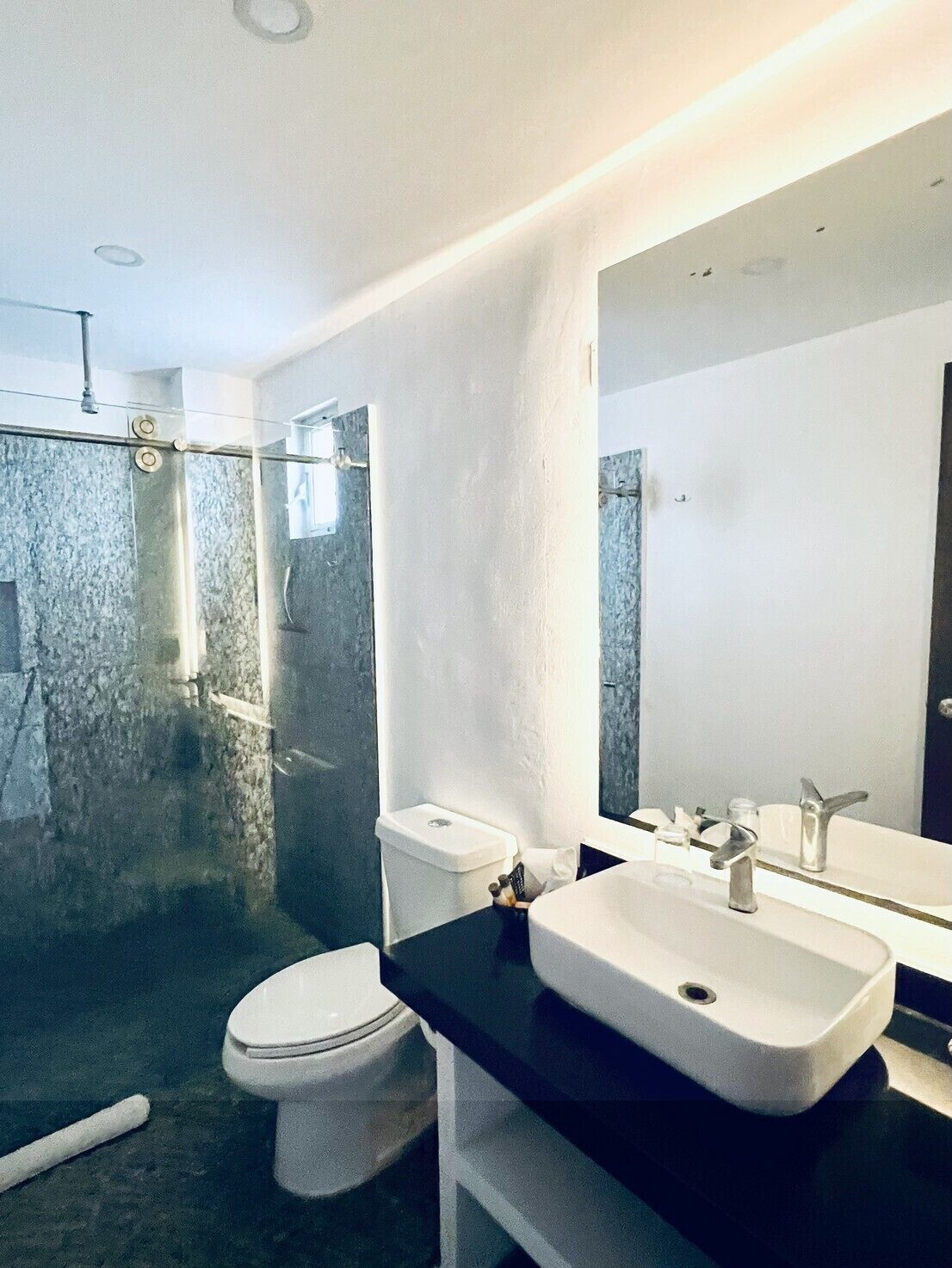 Un baño moderno con ducha de vidrio, inodoro blanco y tocador negro con lavamanos y espejo.