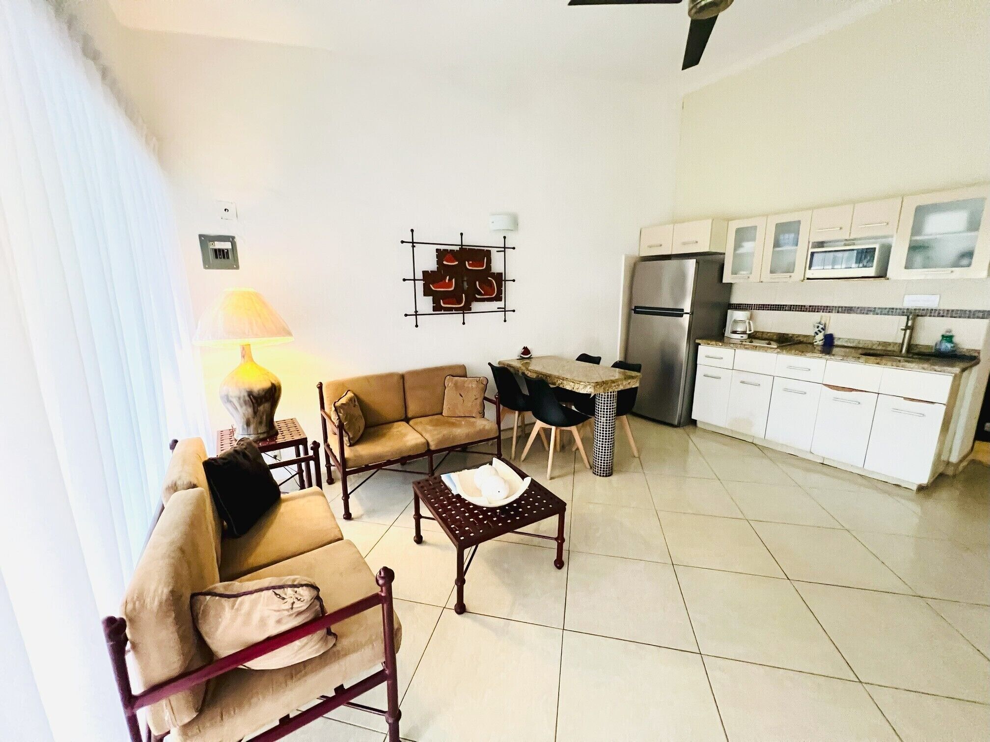 A living room with a sofa, a coffee table, and a dining table with chairs. There is a kitchen area with a refrigerator and cabinets.