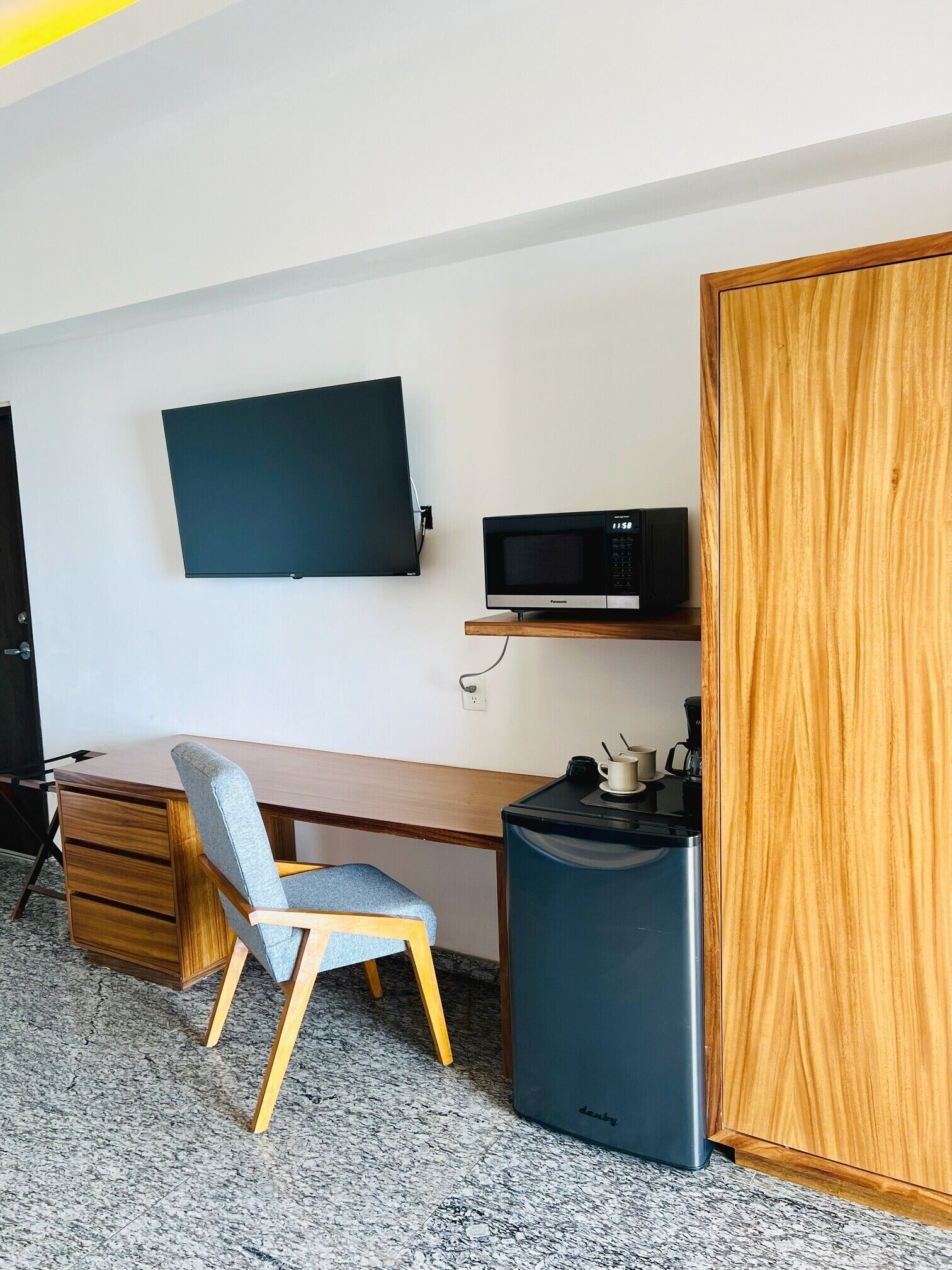 A compact room with a wooden desk, a chair, a flat-screen TV mounted on the wall, a microwave, and a coffee machine.