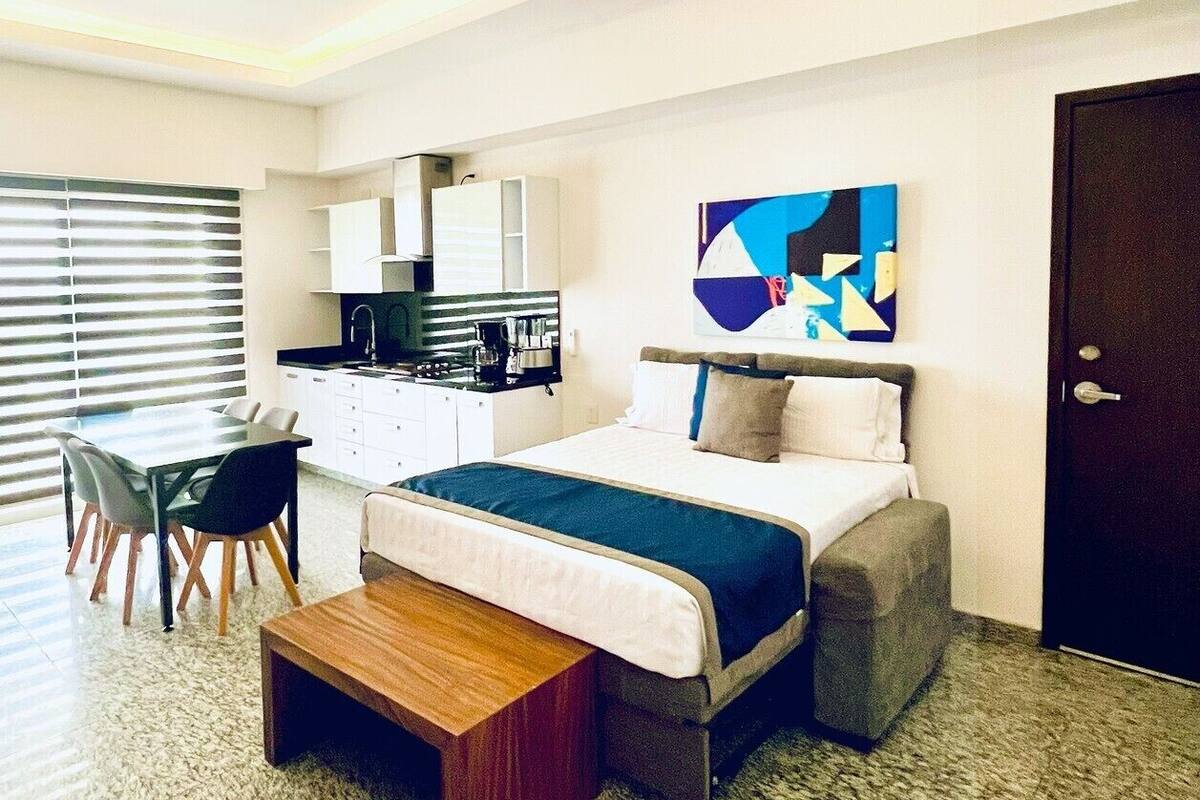 Egy modern szállodai szoba, amelyben található egy ágy, egy íróasztal, egy kis konyhasarok és egy mennyezeti ventilátor.