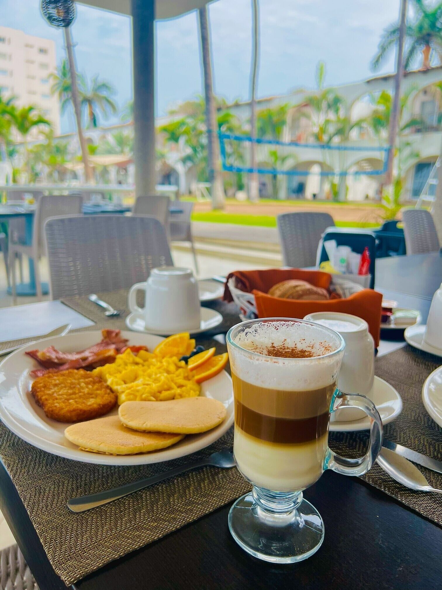 Une table de petit-déjeuner avec une assiette d’œufs, de bacon et de crêpes, un verre contenant une boisson à plusieurs couches, et une vue sur les palmiers et les bâtiments à l’extérieur.