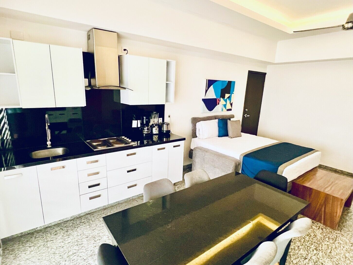 A compact hotel room with a kitchenette, a bed with a blue accent, and a small dining table.