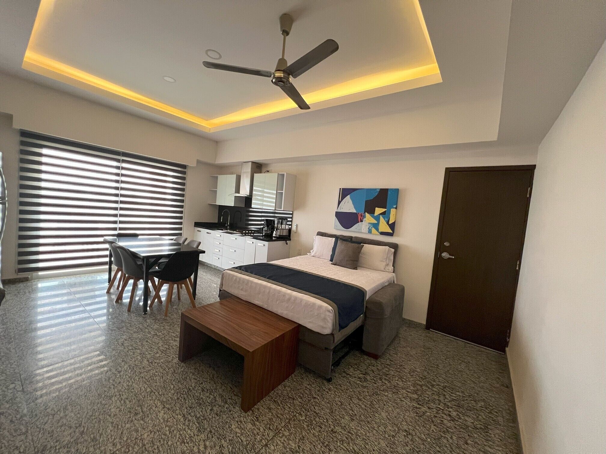 A modern hotel room with a bed, desk, and chair, a ceiling fan, and a painting on the wall.