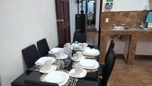 On aperçoit une salle à manger avec une table dressée pour six personnes, comprenant des assiettes, des tasses et des couverts. À l’arrière-plan, on distingue un coin cuisine avec un évier et un plan de travail.