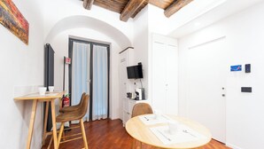 A modern, well-lit room with a wooden floor, a round table with a chair, a small bar area with stools, a television mounted on the wall, and a door with a fire extinguisher. - House in the heart of Naples: comfort, style and perfect location (Napoli)