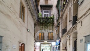 A narrow alleyway with buildings on either side, featuring balconies with plants, a shop with a heart-shaped sign, and a visible entrance to a store. - House in the heart of Naples: comfort, style and perfect location (Napoli)