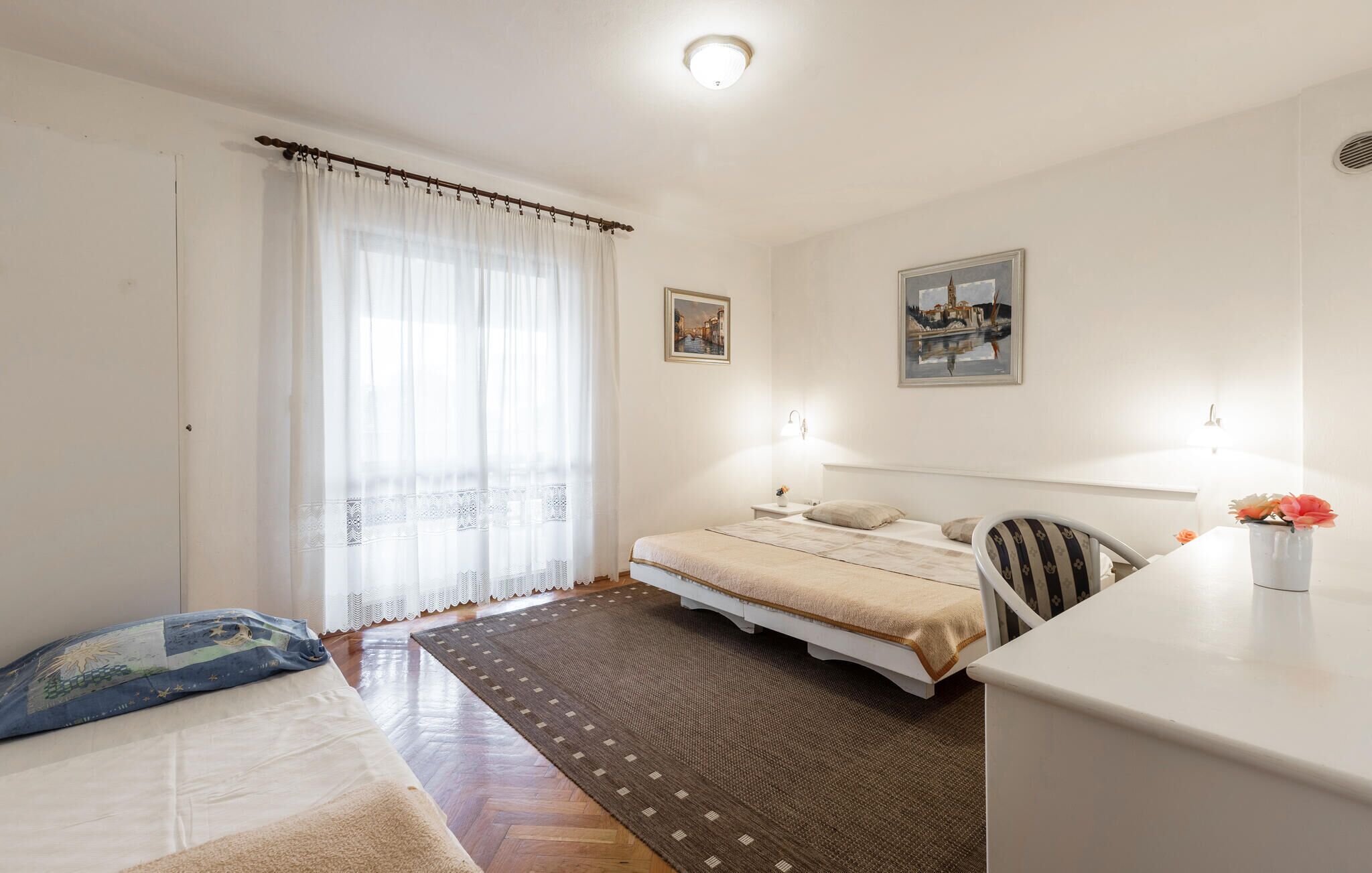 A bedroom with a bed, a desk, a chair, a window with curtains, and two wall-mounted lights.