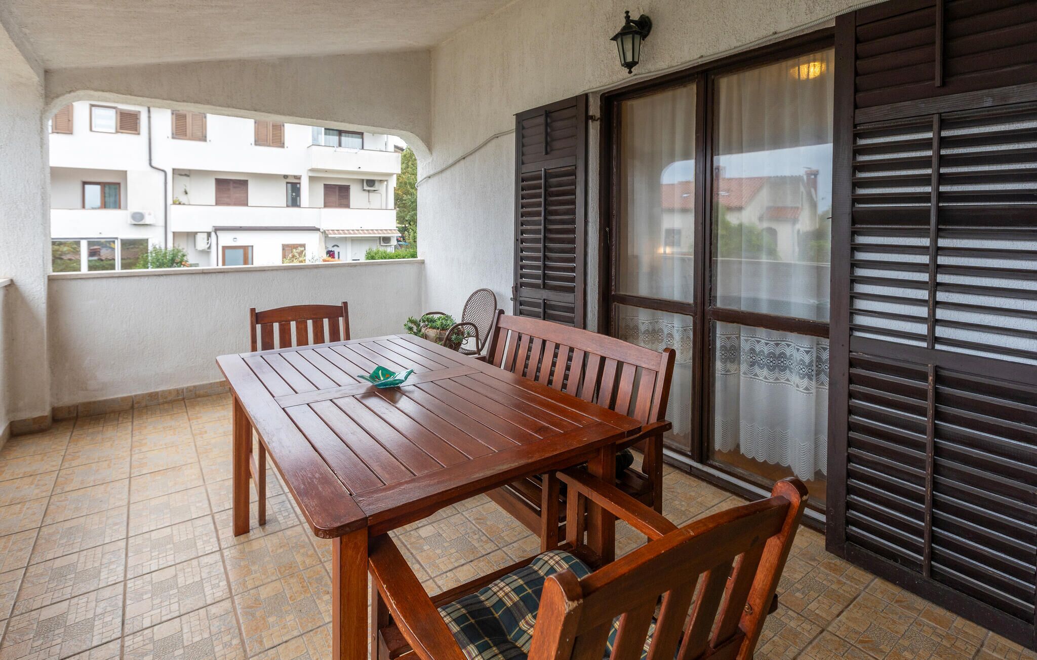 Een balkon met een houten tafel en stoelen, uitzicht op een woongebouw en een raam met zonwering.