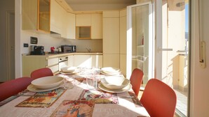 Zona pranzo con tavolo apparecchiato per quattro, cucina sullo sfondo e vista attraverso una porta in vetro.