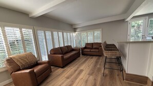 A spacious living room with a brown leather sofa, a smaller sofa, and a wicker chair. Large windows with blinds allow natural light.