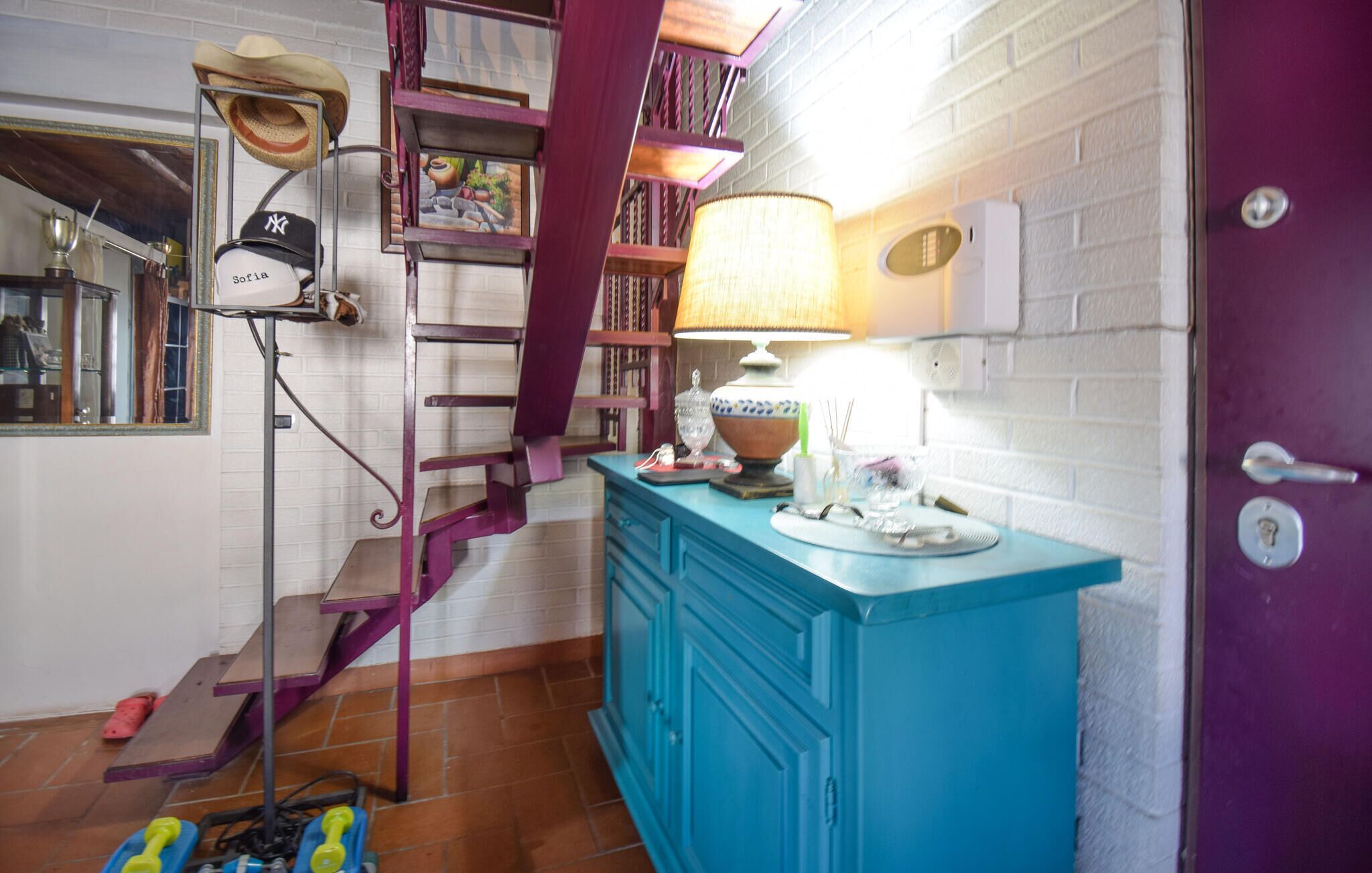 A room with a purple door, a blue cabinet, a staircase with purple railings, and a mirror with a hat on it.