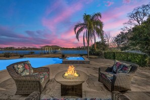 Un patio al aire libre con mobiliario de mimbre y una chimenea, con vistas a una piscina y una masa de agua.