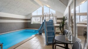 Une piscine intérieure avec un toboggan bleu, une table entourée de chaises et une vue sur l’extérieur grâce à de grandes fenêtres.
