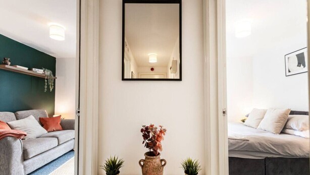 Ett modernt vardagsrum med en grå soffa, ett soffbord och en vas med torkade blommor. Det finns en spegel på väggen och en liten krukväxt på bordet.