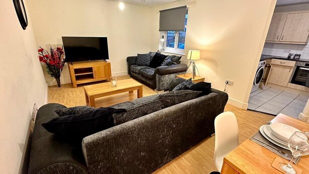 Ein Wohnzimmer mit einem dunkelgrauen Sofa, einem Flachbildfernseher, einem Couchtisch und einer Essecke mit Tisch für zwei Personen.