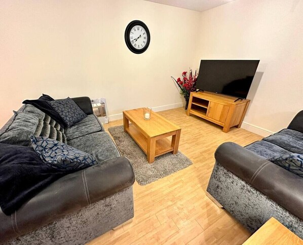 Una sala de estar con dos sofás grises, una mesa de centro de madera, un mueble de televisión con un televisor de pantalla plana y un reloj de pared.
