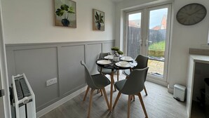 A dining area with a round table, four chairs, a pendant light, two wall-mounted paintings, a clock, and a sliding glass door leading to an outdoor area. - Bolehyde House Long Stay Discounts Free Parking (Swindon)