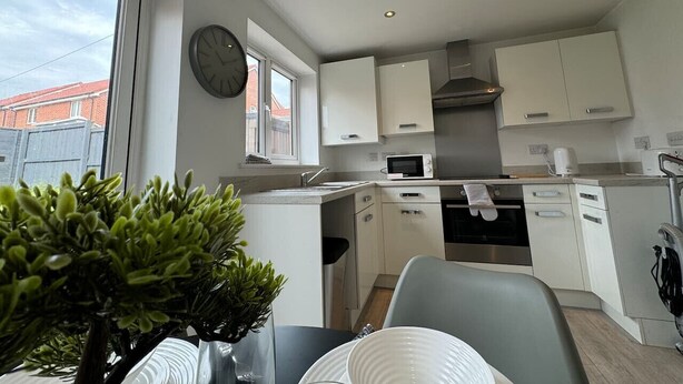 A modern kitchen with white cabinets, a microwave, and a sink. There is a clock on the wall and a potted plant on the table. - Bolehyde House Long Stay Discounts Free Parking (Swindon)