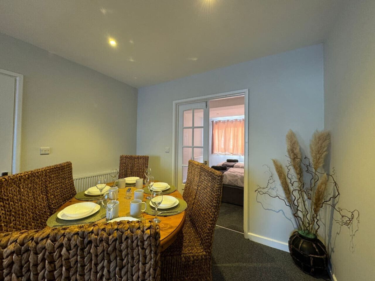 A dining area with a round table set for four, surrounded by wicker chairs. There is a vase with pampas grass on the wall.