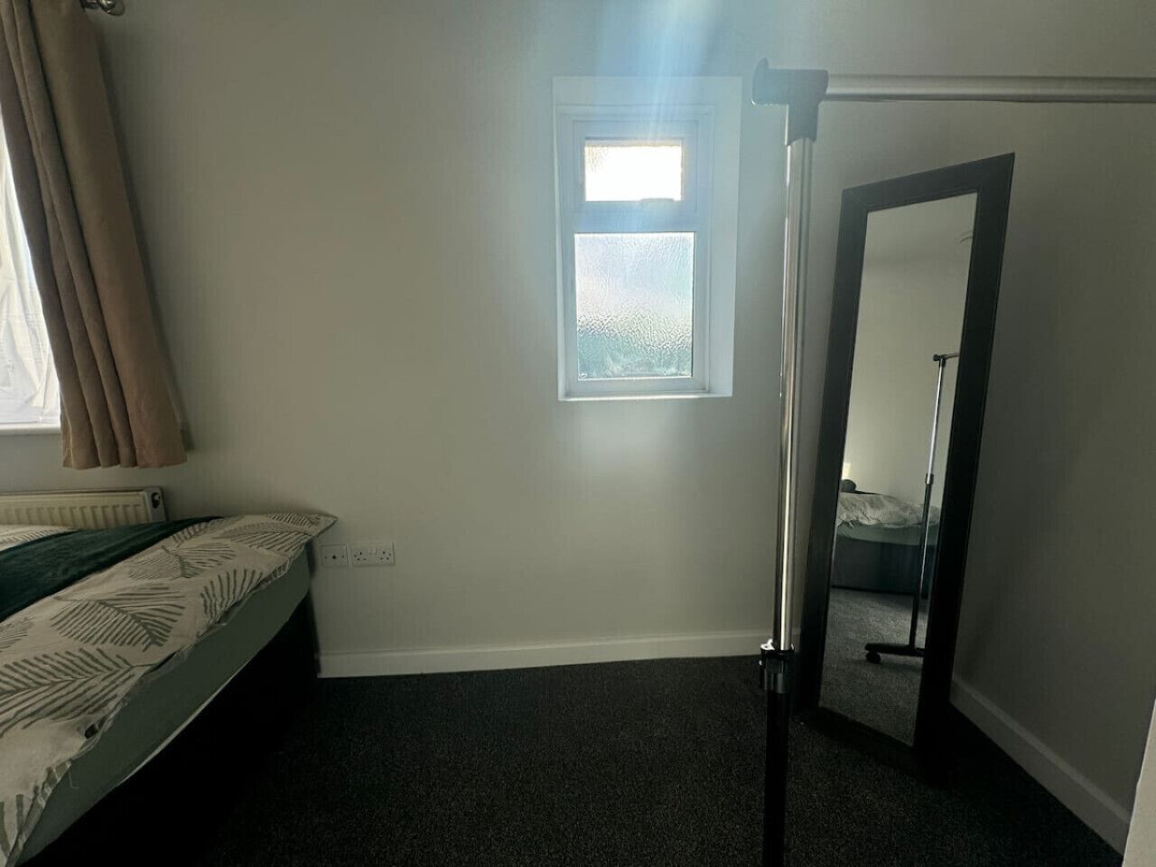 A small, sparsely furnished room with a single bed, a window, and a door leading to another room.