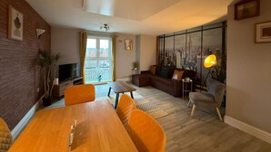 A modern living room with a wooden dining table, orange chairs, a sofa, and a cityscape mural on the wall.