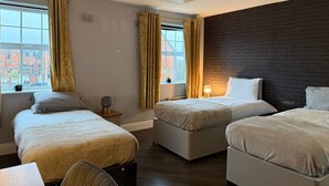 A hotel room with two beds, a window with curtains, a brick accent wall, and a small table with a lamp.