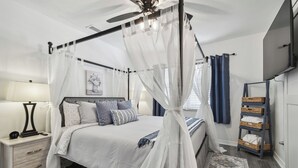 Un dormitorio con cama con dosel, mesita de noche, lámpara, ventilador de techo y televisor montado en la pared.